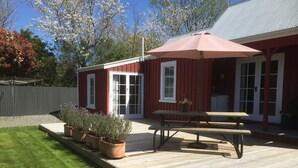 En träveranda med ett picknickbord och ett stort parasoll, omgiven av en röd byggnad med vita detaljer och en täckt uteplats.