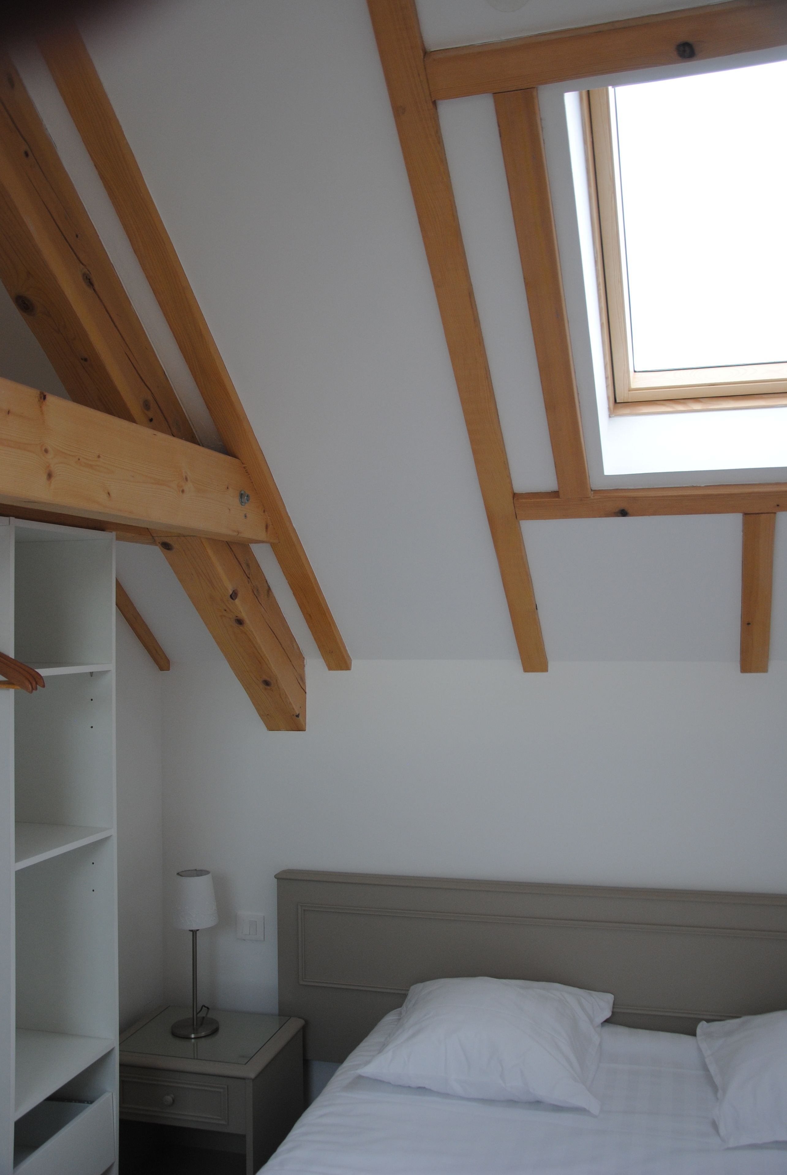 A bedroom with a sloped ceiling, a skylight, a bed with white bedding, a bedside table with a lamp, and a wooden wardrobe.