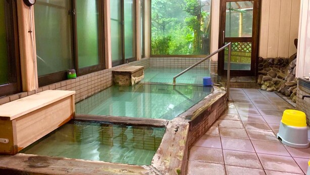 A traditional Japanese onsen with a wooden bench, a stone platform, and a view of greenery outside. - Kirishimaya Ryokan (Kusatsu)
