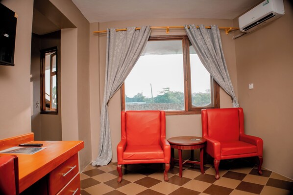 A room with two red chairs, a small round table, a wooden desk, and a window with curtains. - Serene Hotel (Dar es Salaam)