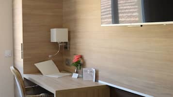 Quarto de hotel compacto com escrivaninha de madeira, uma cadeira, um banquinho, TV fixada na parede e uma mesinha com um vaso de flores.