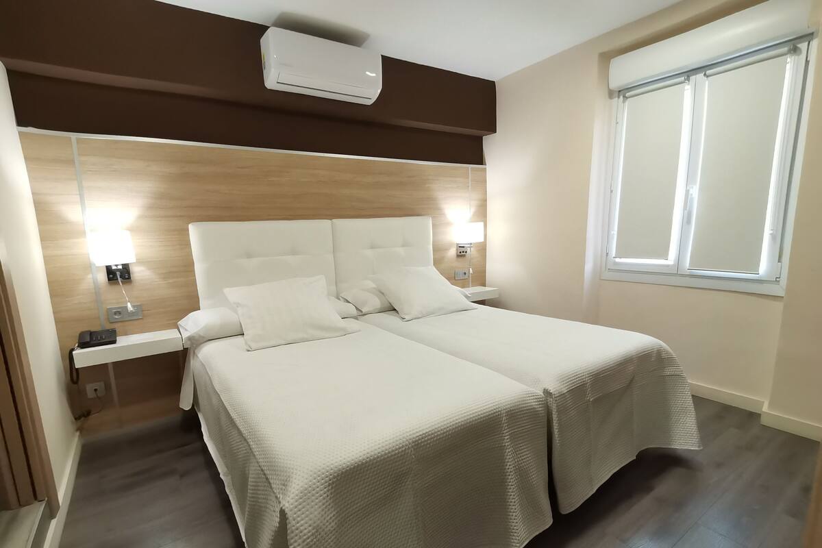 A hotel room with a double bed, bedside lamps, a phone on the nightstand, and a window with blinds.
