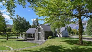 A stone building with a covered porch, surrounded by well-maintained grass and trees. - Taramea, family friendly home walking distance to Mora Wines (Lake Hayes)