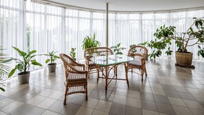 A room with a glass-top table surrounded by wicker chairs, potted plants, and large windows with sheer curtains. - Hotel Galaktika (Lviv)