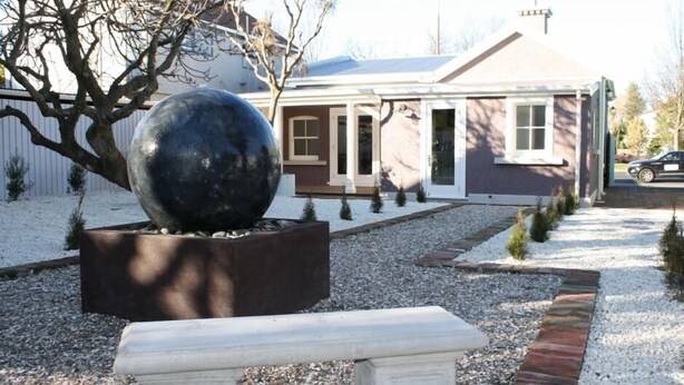 Une maison avec une allée en gravier, une grande sculpture sphérique dans un bac à fleurs et un banc au premier plan.