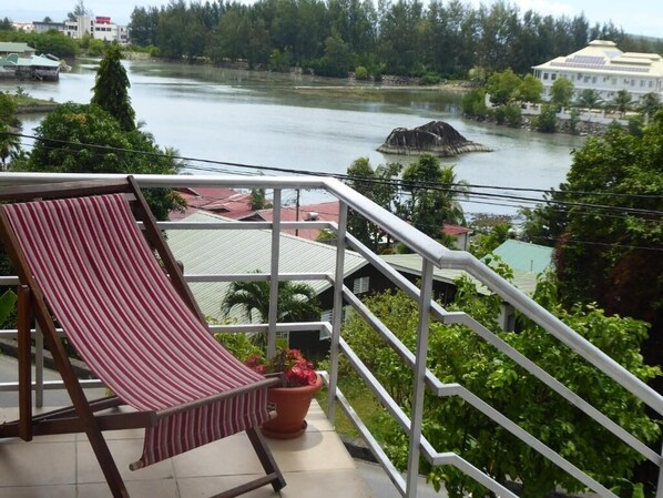 A balcony with a striped chair and a potted plant, overlooking a river with buildings and greenery in the background. - Chez Odet (Mahé Island)