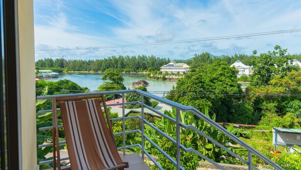 A balcony with a wooden chair, overlooking a river, greenery, and a distant building. - Chez Odet (Mahé Island)