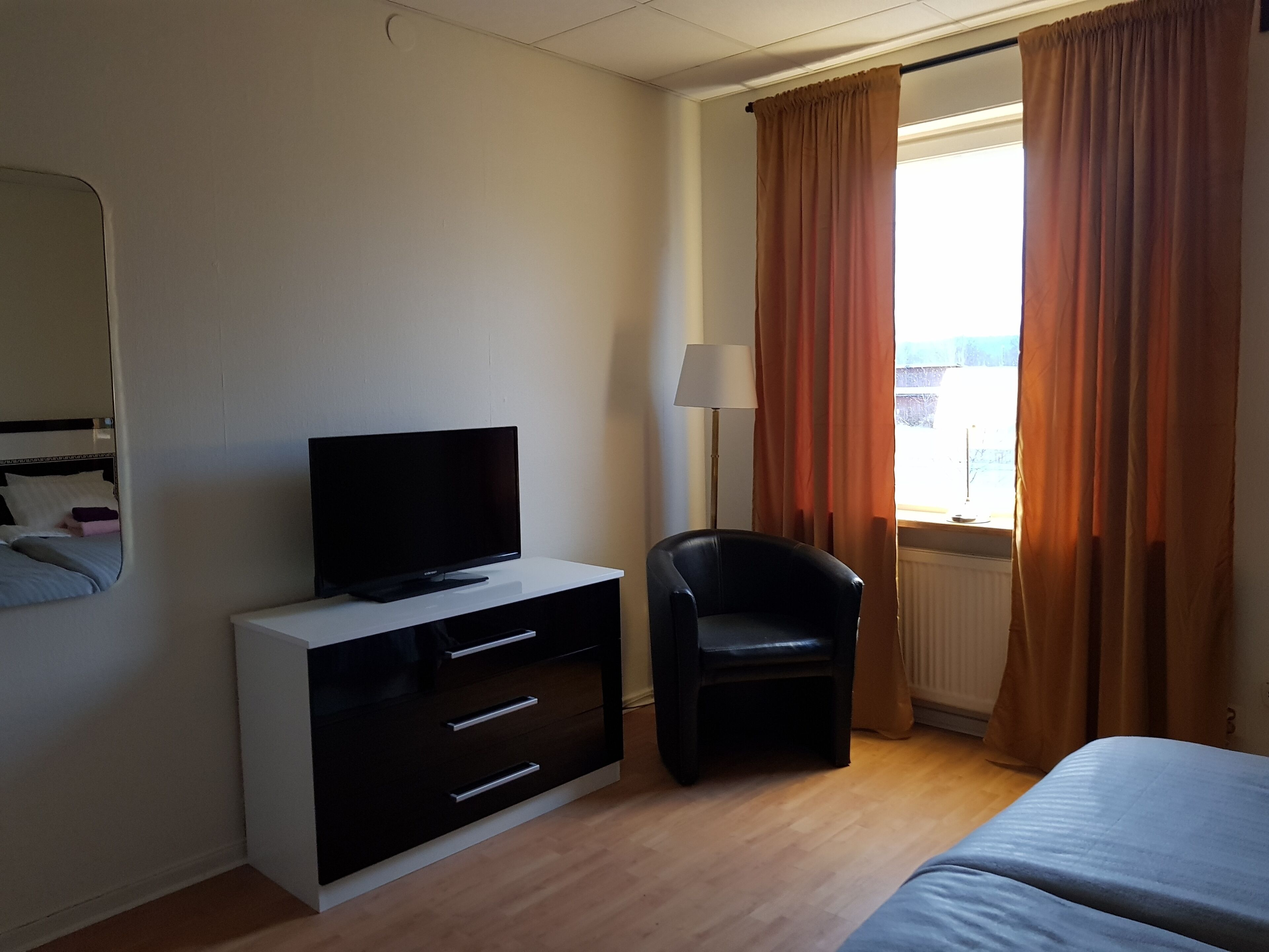Una habitación de hotel con una televisión de pantalla plana sobre una cómoda blanca, una silla negra, una cama y un ventanal con cortinas.