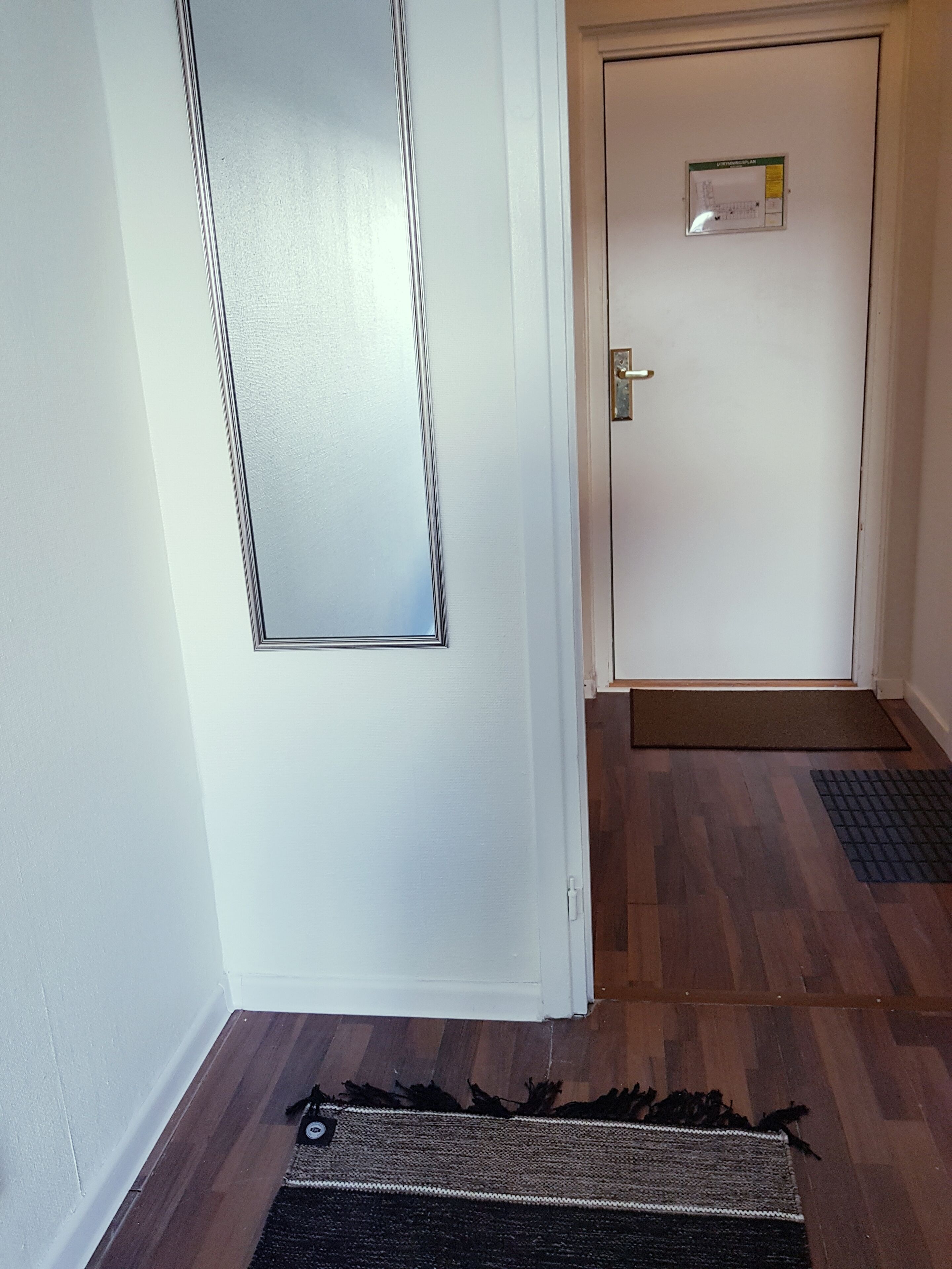 Un couloir avec un plancher en bois, un paillasson et une porte sur laquelle est apposée une pancarte.