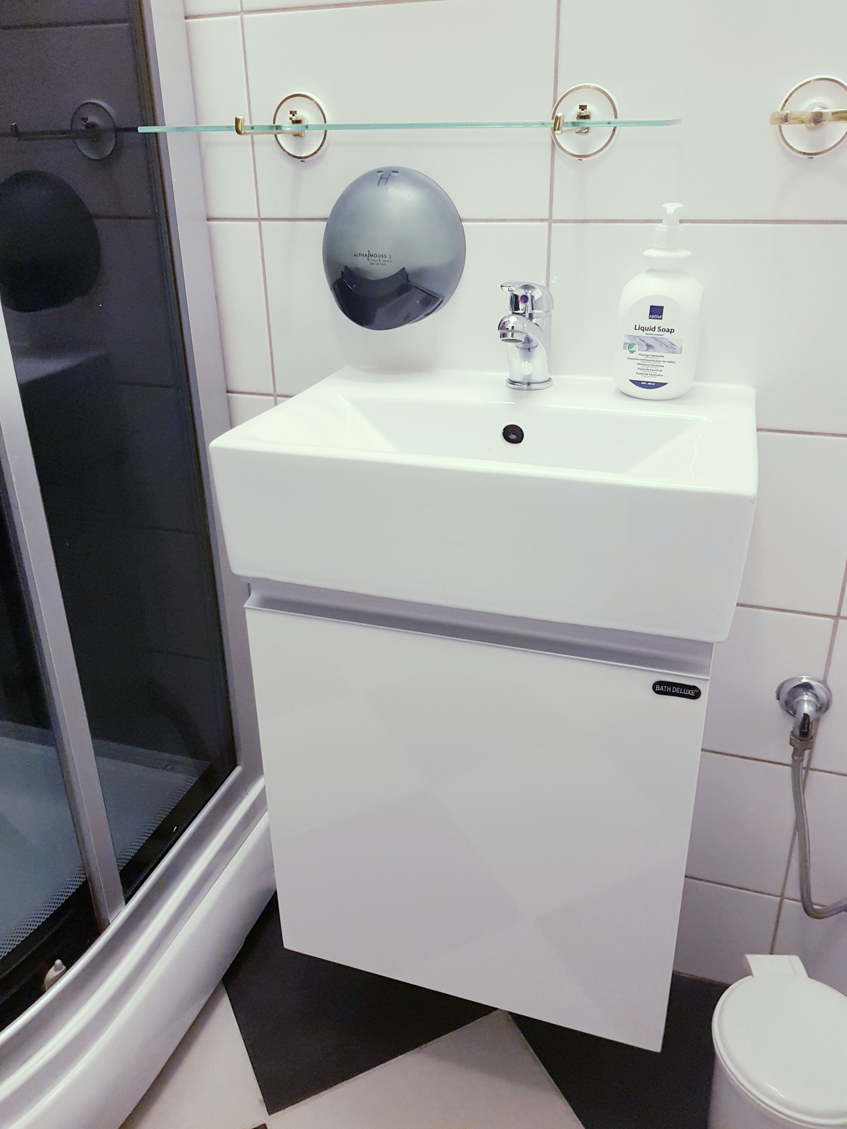 Un baño con un lavamanos blanco, un espejo, estante de vidrio, ducha y retrete.