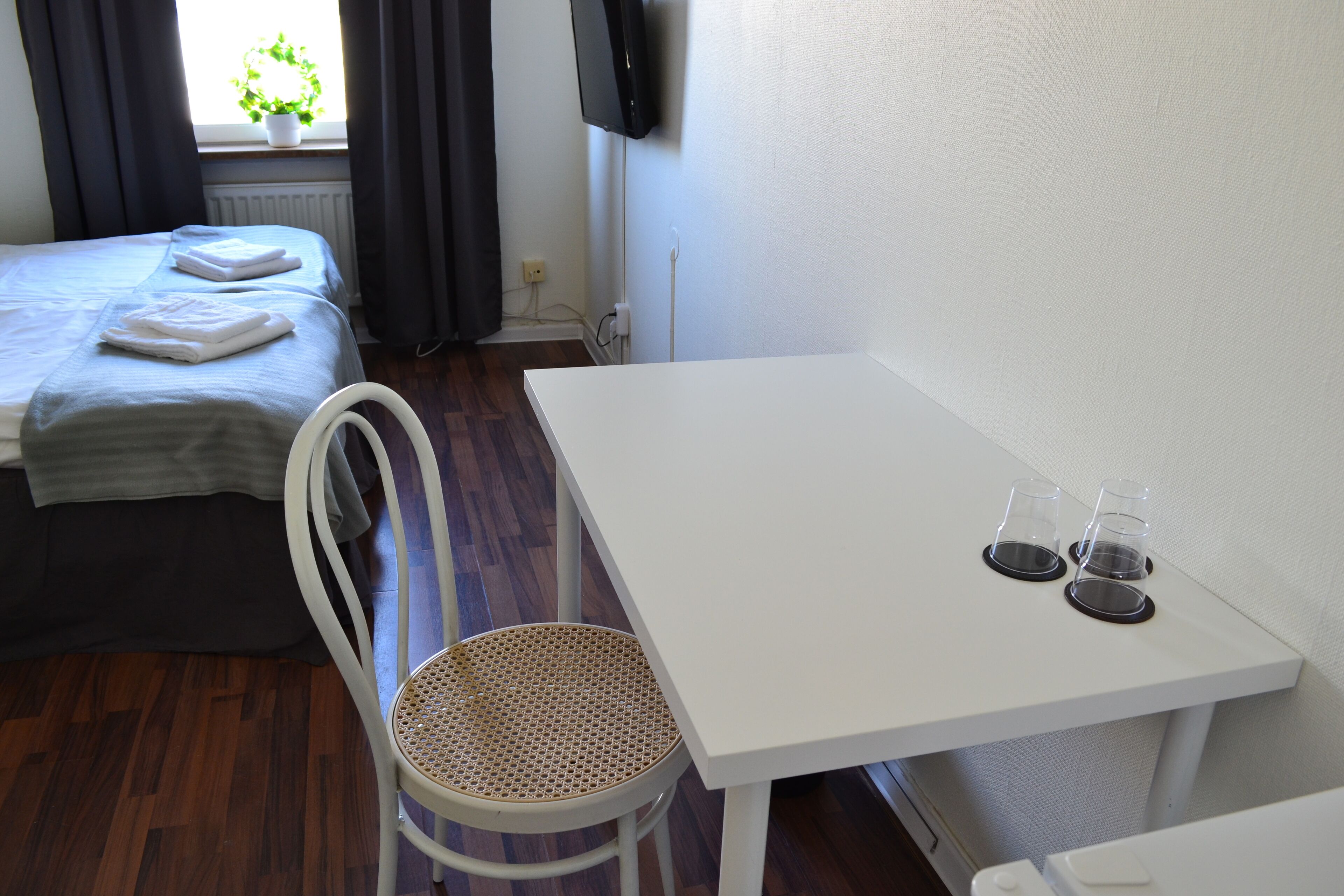 Una camera d'albergo con un letto, un tavolino bianco con due bicchieri, una sedia in vimini e una pianta in vaso vicino alla finestra.