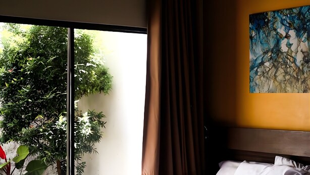 Una cama bien hecha con sábanas y toallas blancas, un ventanal con vistas a la vegetación y un cuadro en la pared.