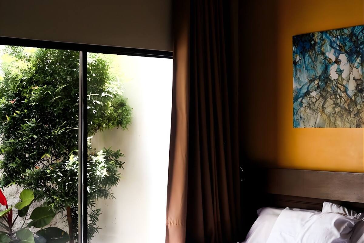 Una cama bien hecha con sábanas y toallas blancas, un ventanal con vistas a la vegetación y un cuadro en la pared.