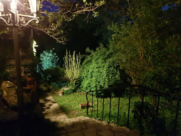 Ein Gartenpfad bei Nacht mit einer Steinsitzbank, einem kleinen Baum und einem Metallzaun.