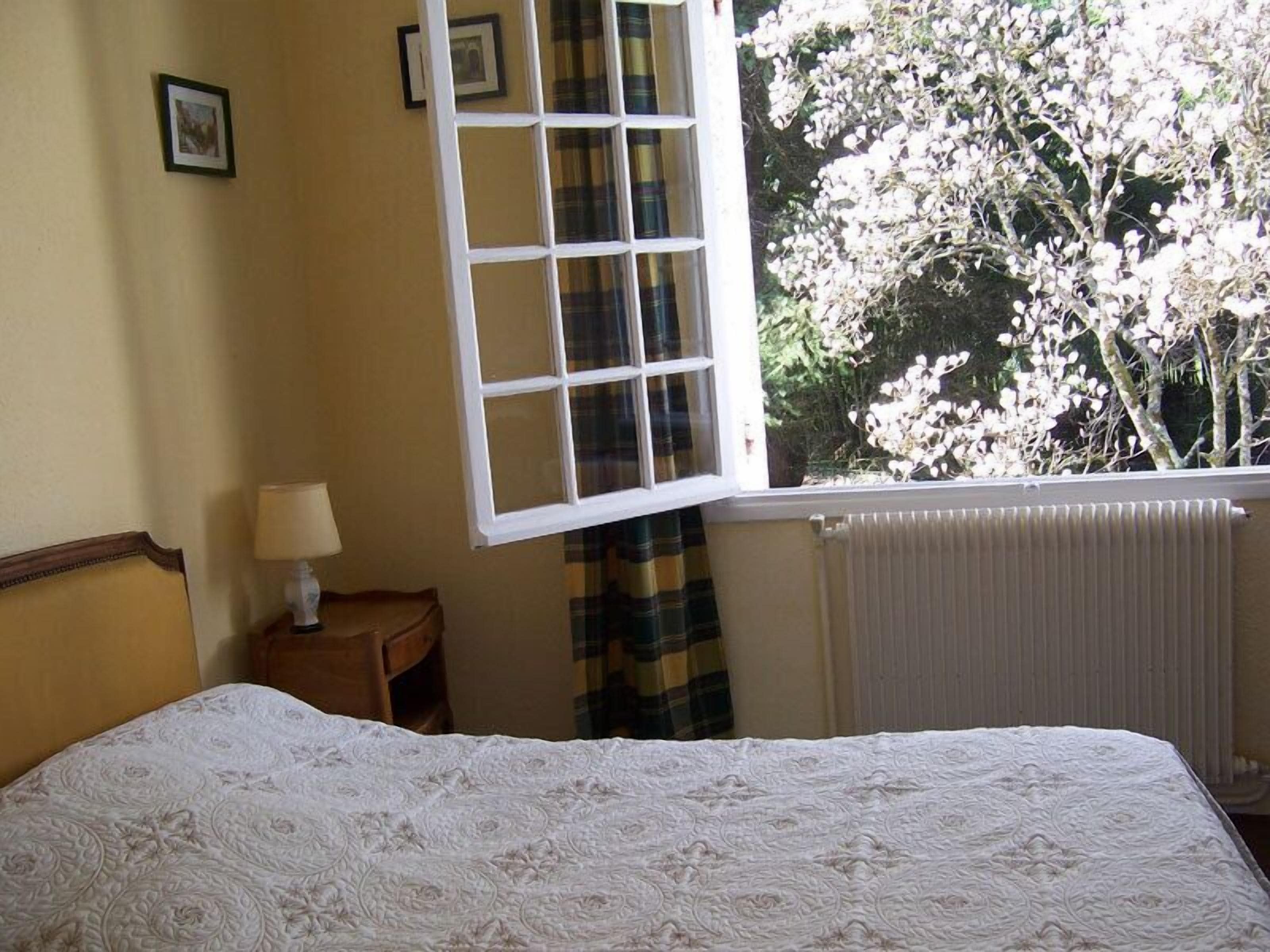 A bedroom with a bed, a nightstand, a lamp, a window with a view of trees, and a radiator.