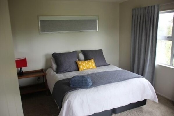 A neatly made bed with a white and grey duvet, two pillows, and a blue blanket. There is a red lamp on a wooden bedside table.