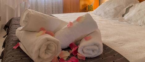 A neatly made bed with rolled towels and rose petals, a bedside table with a lamp, and a window with curtains.