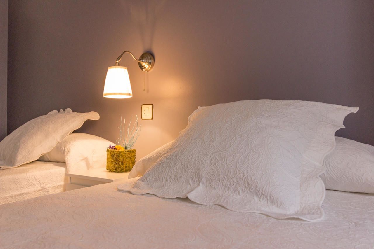 Een net opgemaakt bed met witte lakens en kussens, een nachtkastje met een lamp en een kleine vaas met gedroogde bloemen.