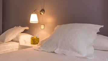 Een net opgemaakt bed met witte lakens en kussens, een nachtkastje met een lamp en een kleine vaas met gedroogde bloemen.