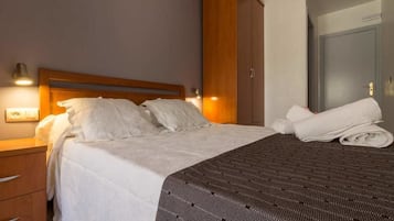 Una cama bien hecha con sábanas blancas y una manta oscura estampada, cabecero de madera y una lámpara fijada a la pared.
