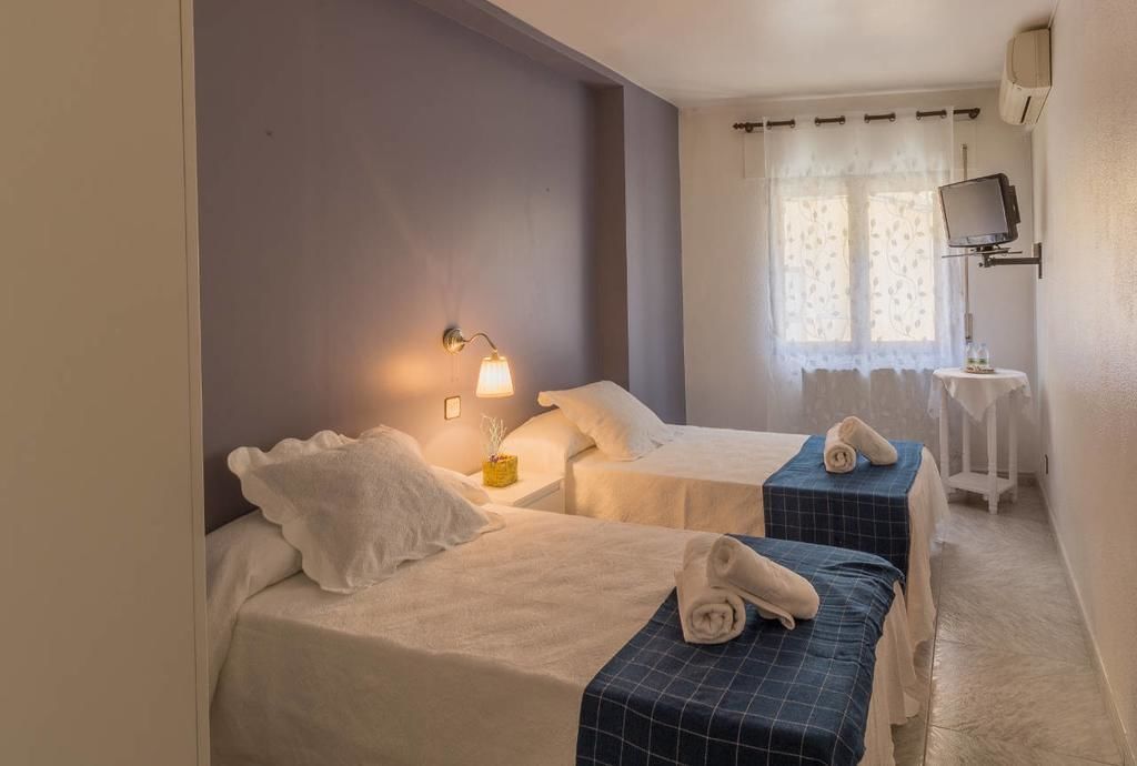 Habitación de hotel con dos camas, una mesita con lámpara, un televisor en un mueble bajo y una ventana con cortinas.