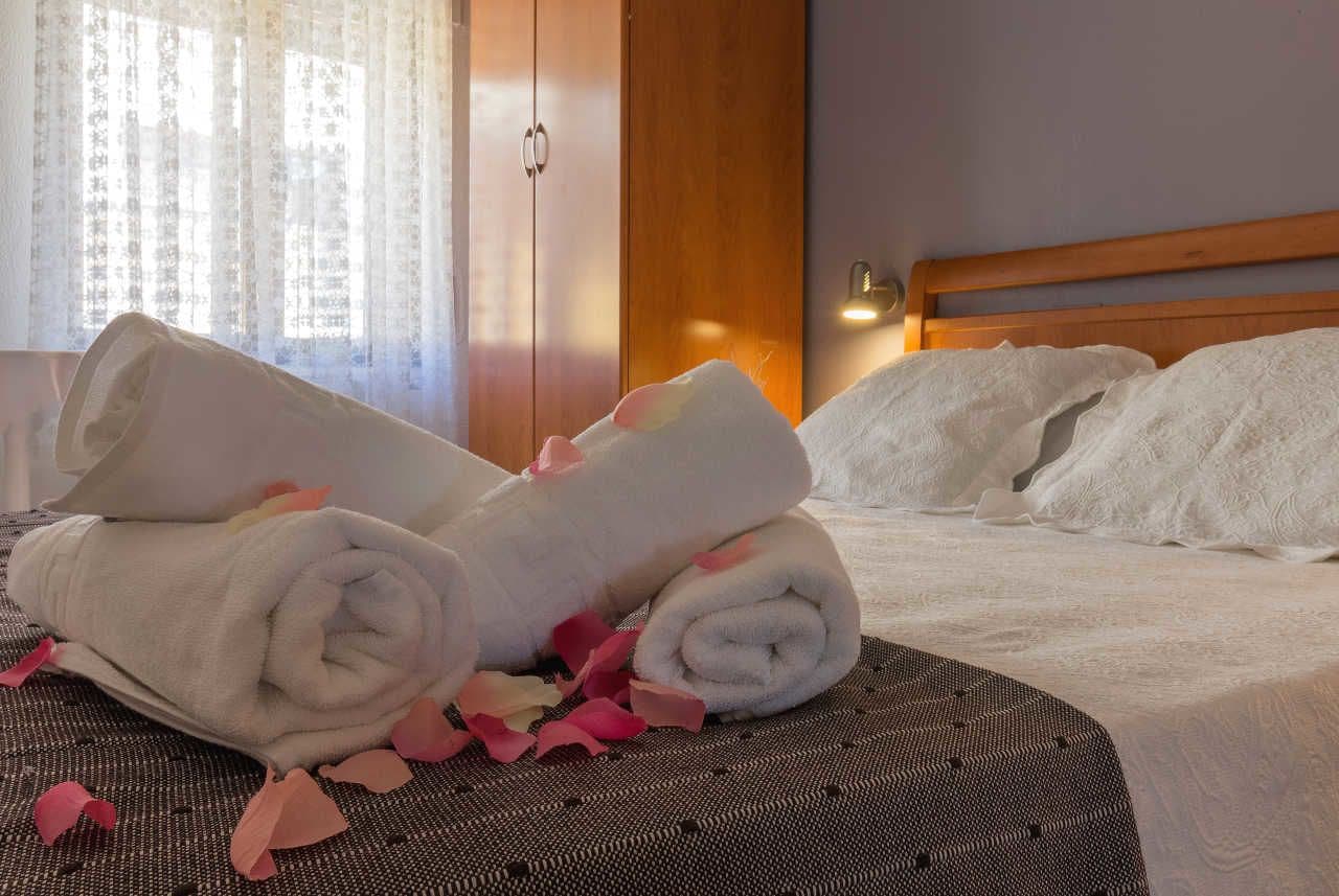 Una cama bien hecha con toallas blancas enrolladas y pétalos de rosa, un cabecero de madera y un armario al fondo.