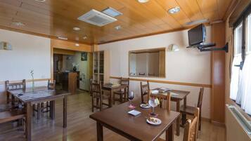A dining area with wooden tables and chairs, a television mounted on the wall, and a window providing natural light.