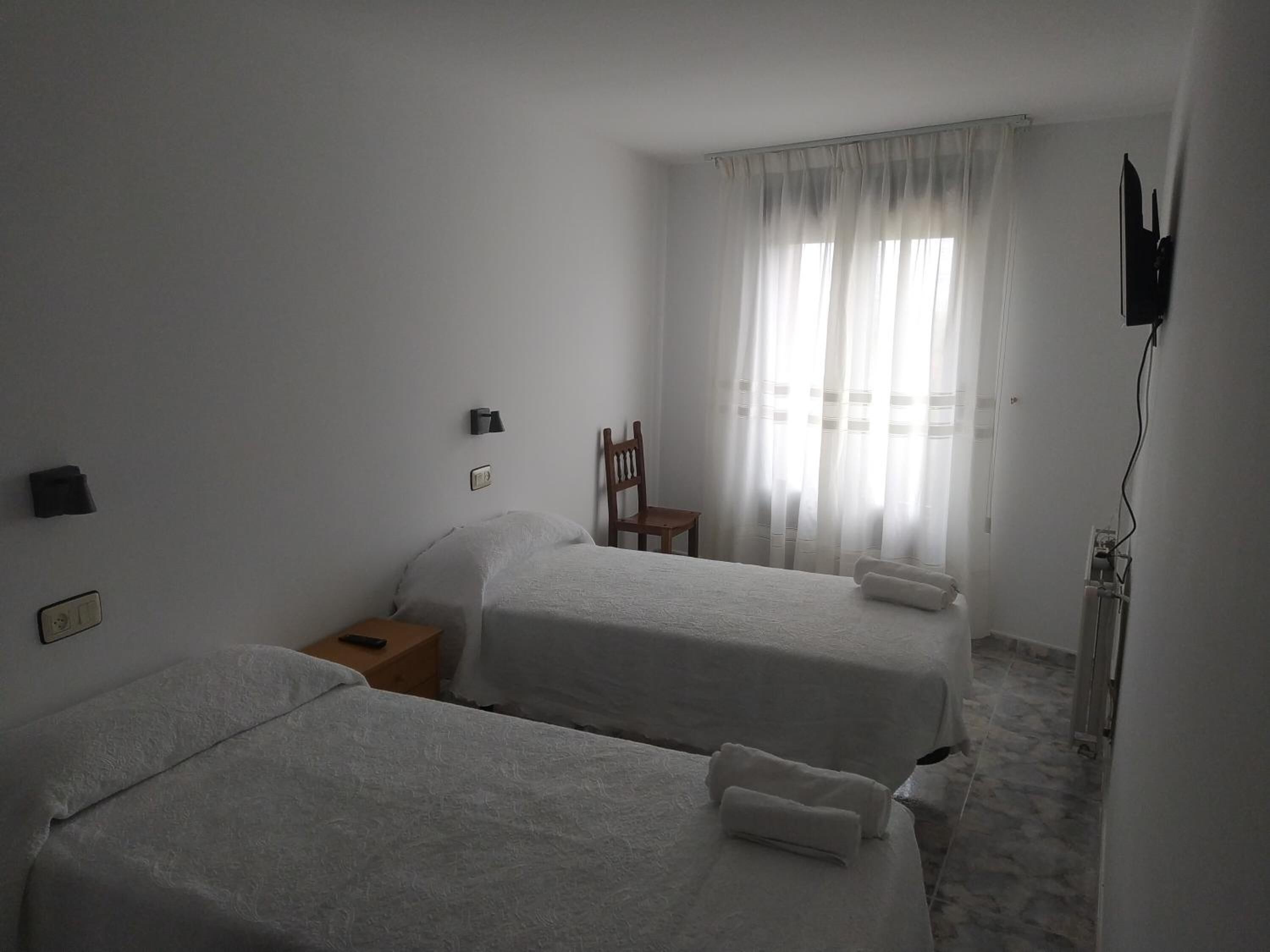 A hotel room with two beds, a chair, a nightstand, a TV, and a window with sheer curtains.