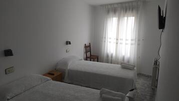 A hotel room with two beds, a chair, a nightstand, a TV, and a window with sheer curtains.