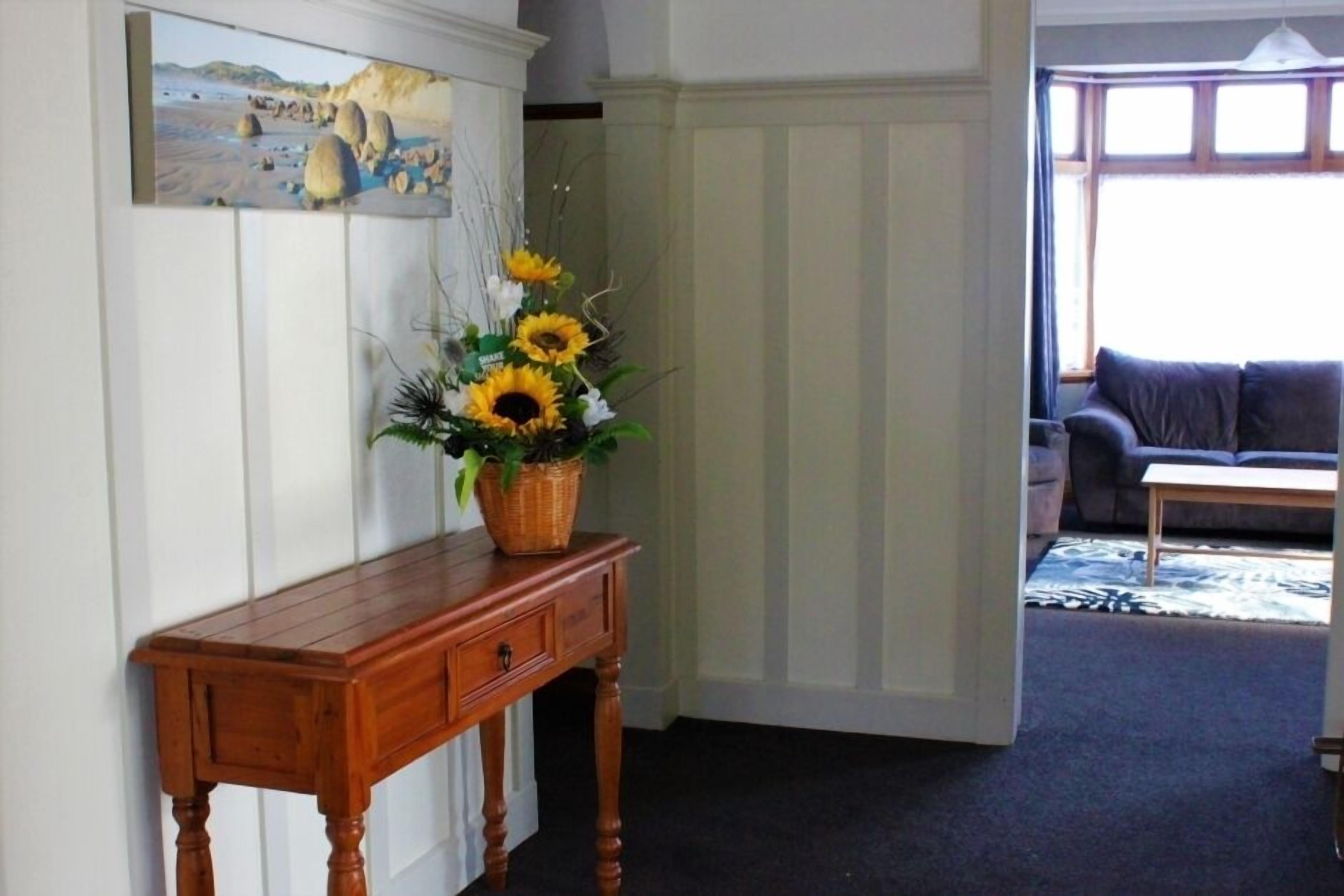 Een houten console tafel met een vaas zonnebloemen, een schilderij van een strandgezicht, en een uitzicht op een woonkamer met een bank en een raam.