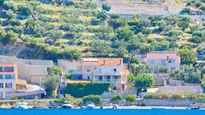 A coastal town with buildings along the waterfront, boats docked, and a hillside with vegetation. - Apartments Violic - Standard Two Bedroom Apartment with Terrace and Sea View (A5) (Zaton)
