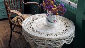 Una habitación con una silla de madera, una mesa redonda con mantel de encaje y un jarrón con flores.
