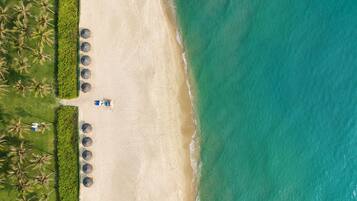 Vista aerea di una spiaggia con una fila di ombrelloni, un vialetto e palme.