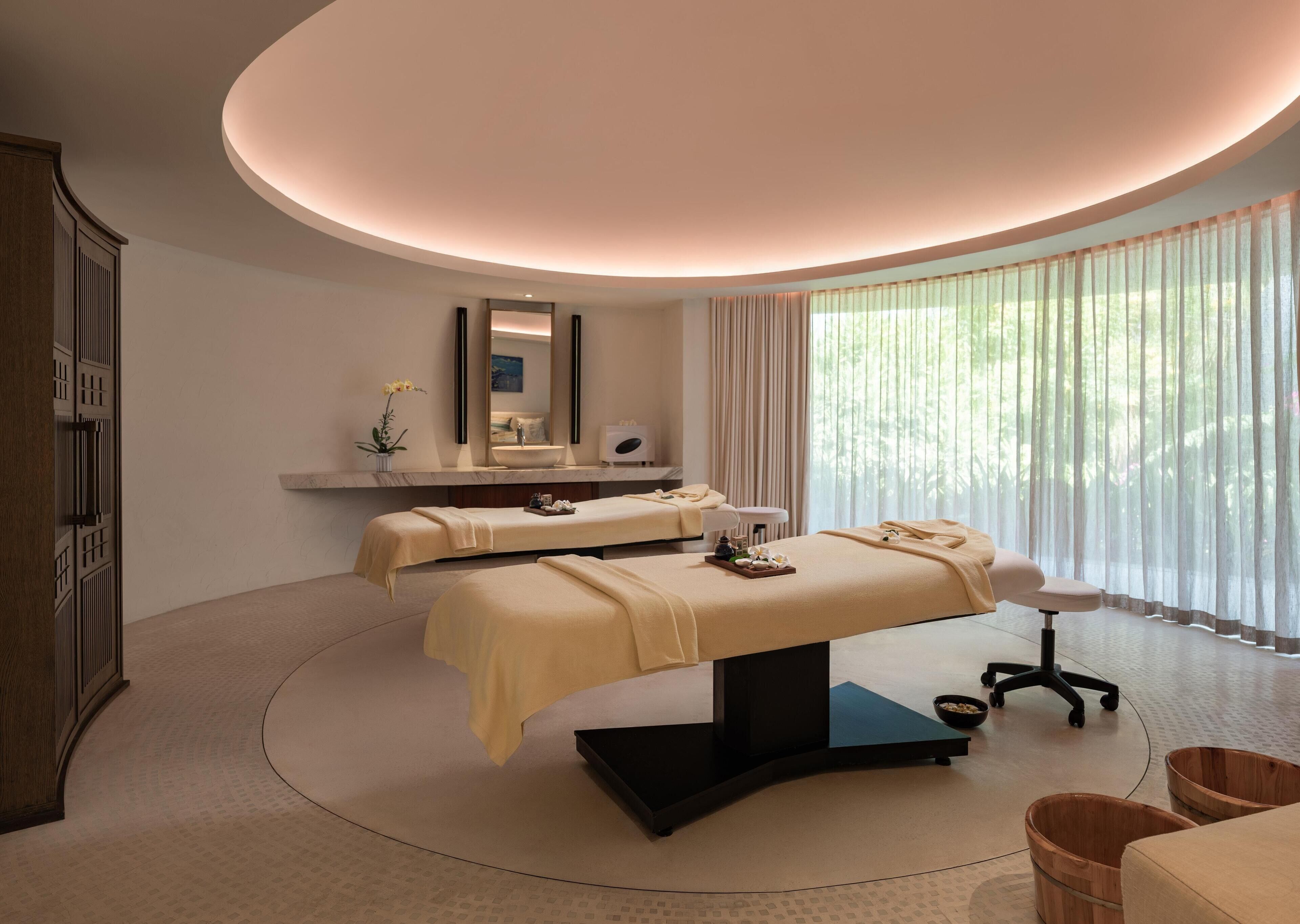 A spa room with two massage tables, a large mirror, and a view of greenery through sheer curtains.