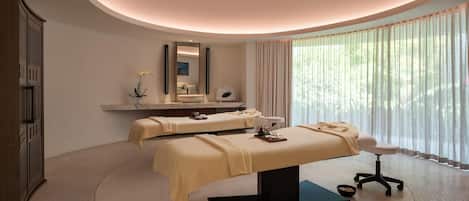 A spa room with two massage tables, a large mirror, and a view of greenery through sheer curtains.