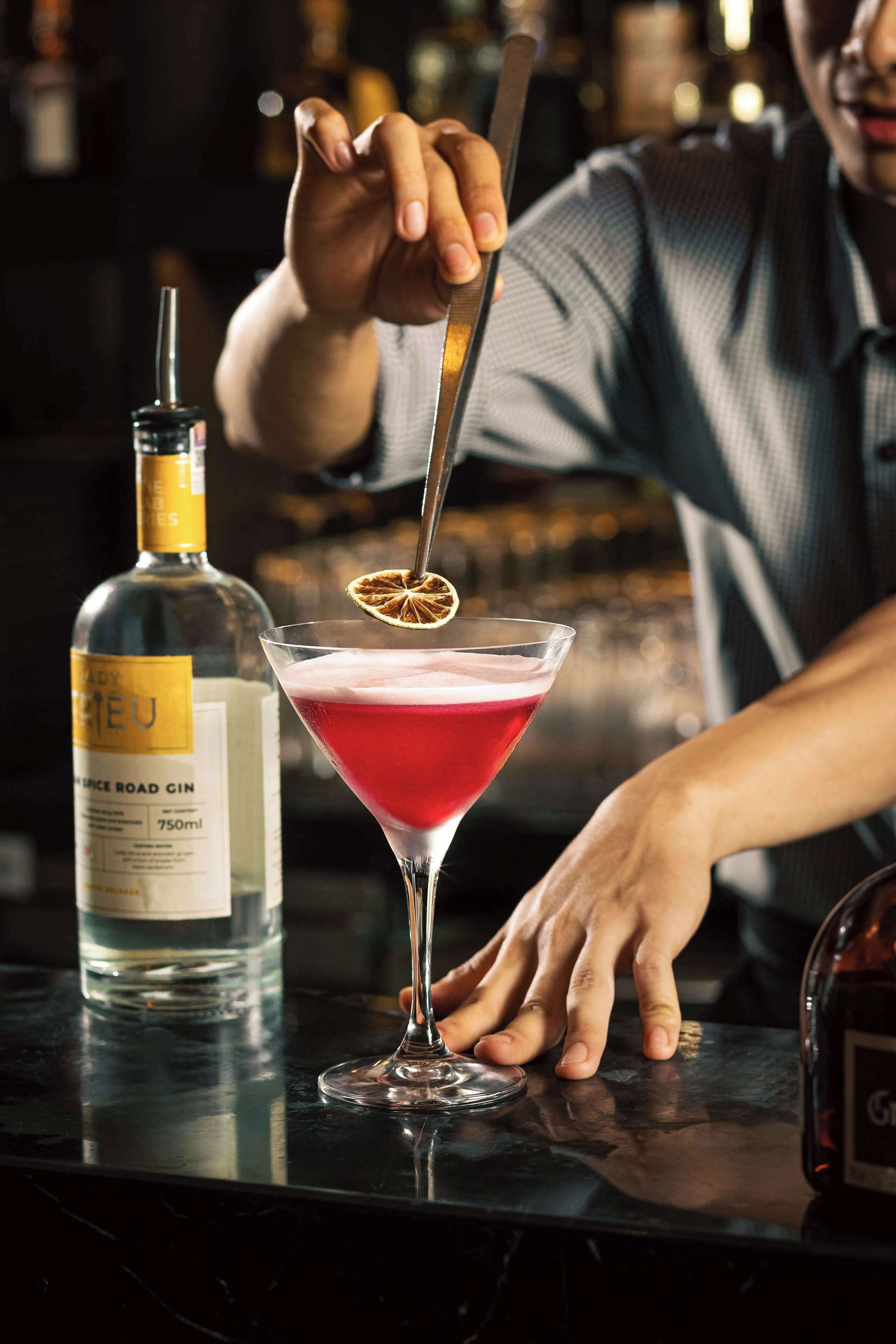 Un barman prépare un cocktail à l’aide d’une bouteille de gin et d’un verre à martini, le tout sur le comptoir du bar.