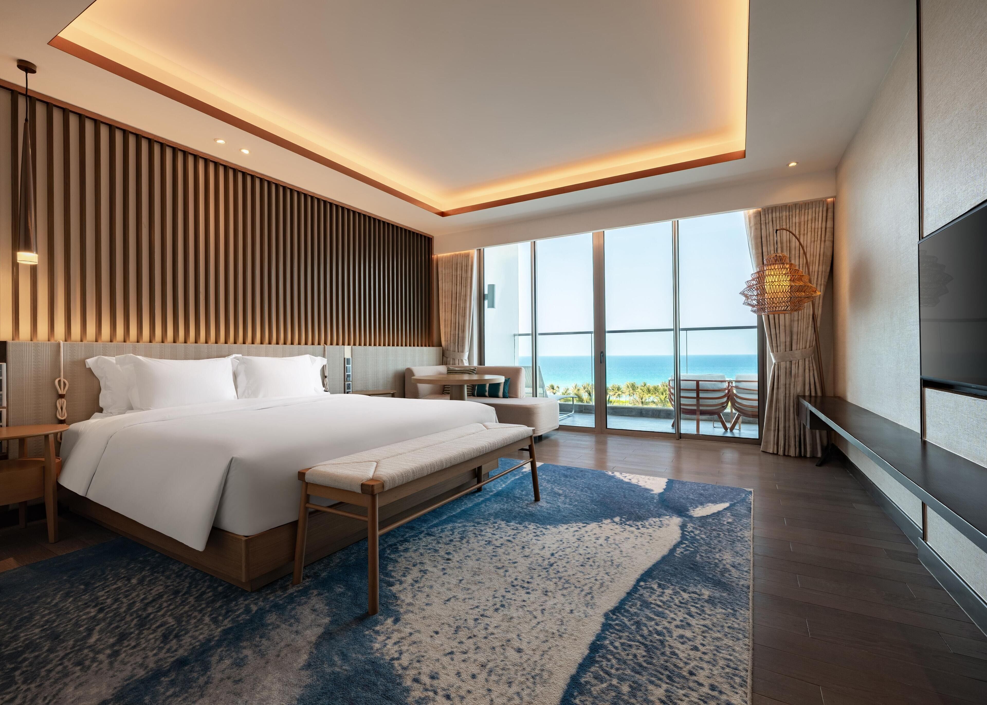 A modern hotel room with a large bed, a seating area, a TV, and a view of the ocean.