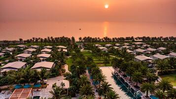 Un resort con varias construcciones tipo bungalow, un área de piscina central y vistas al atardecer sobre el agua.