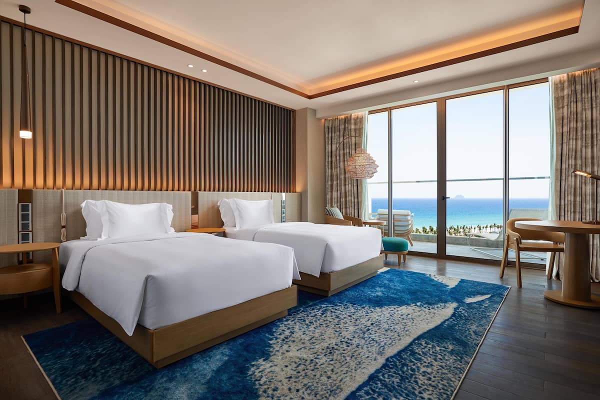 Ein modernes Hotelzimmer mit zwei Betten, einem großen Fenster mit Meerblick, einem blauen Teppich mit Muster und einer Holzbeschichtung der Decke.