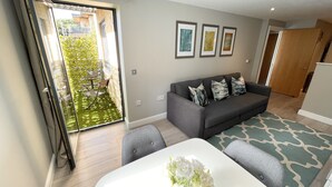 A modern living room with a grey sofa, a white coffee table, and a patterned rug. There are three framed pictures on the wall and a sliding glass door leading to a balcony with a garden. - The Wesley Luxury City Apartment (Oxford)