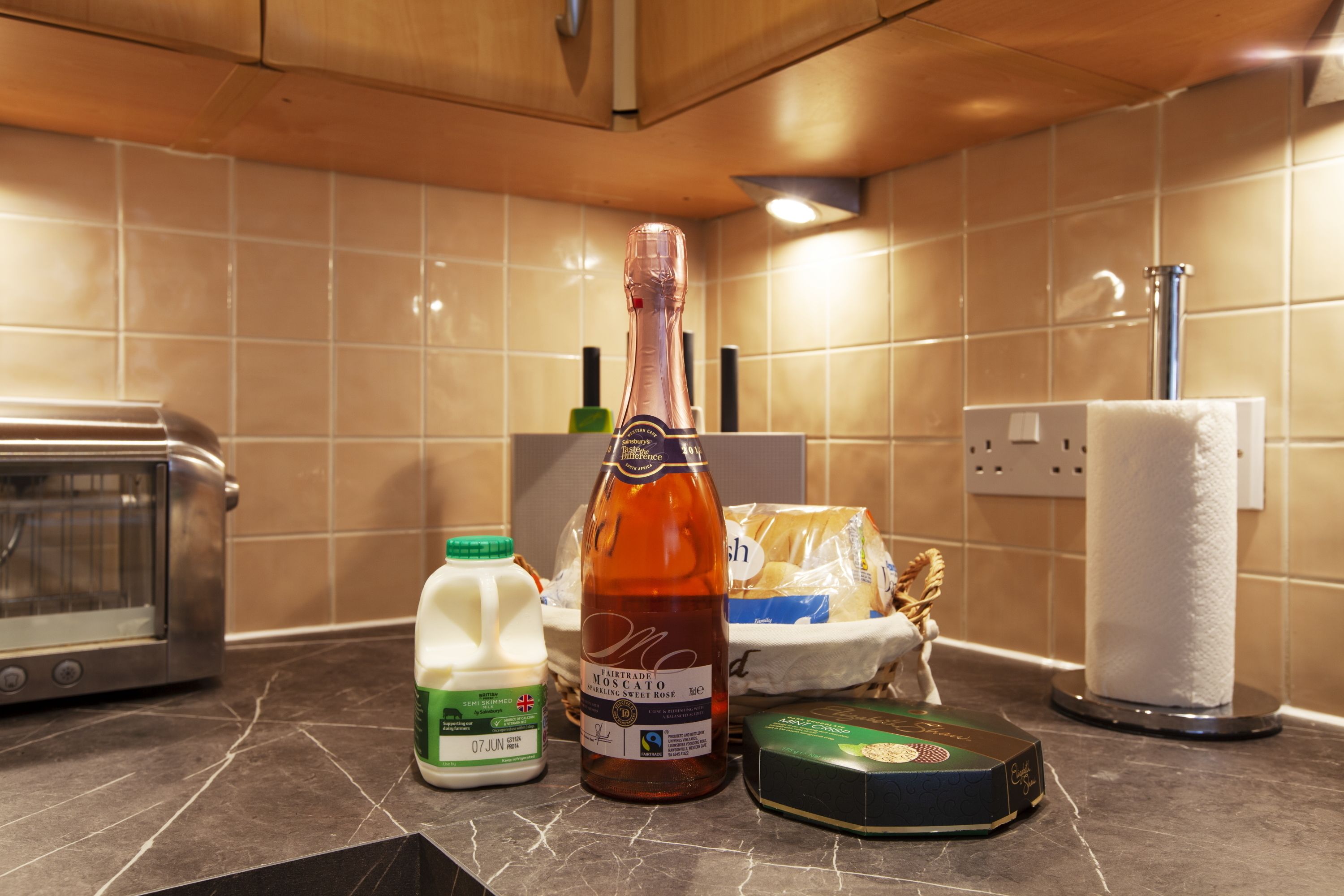 A kitchen counter with a bottle of sparkling Moscato, a carton of milk, a loaf of bread, and a box of cheese.