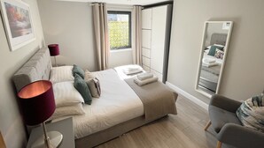 A modern bedroom with a large bed, a bedside lamp, a mirror, and a window with curtains. - The Wesley Luxury City Apartment (Oxford)
