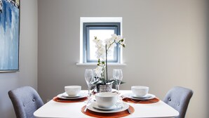 A dining table set with white bowls, plates, and cutlery, flanked by grey upholstered chairs, with a window and a painting in the background. - The Wesley Luxury City Apartment (Oxford)