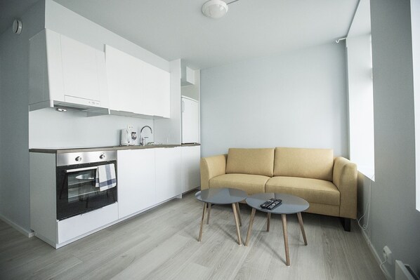 Cocina compacta con armarios blancos, horno empotrado y cafetera. Hay un sofá color beige y dos mesitas redondas con controles remotos.