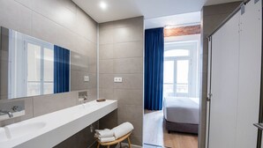 A modern bathroom with a large mirror, a white sink, and a wooden stool with towels. -  Room Gran Vía (Madrid)