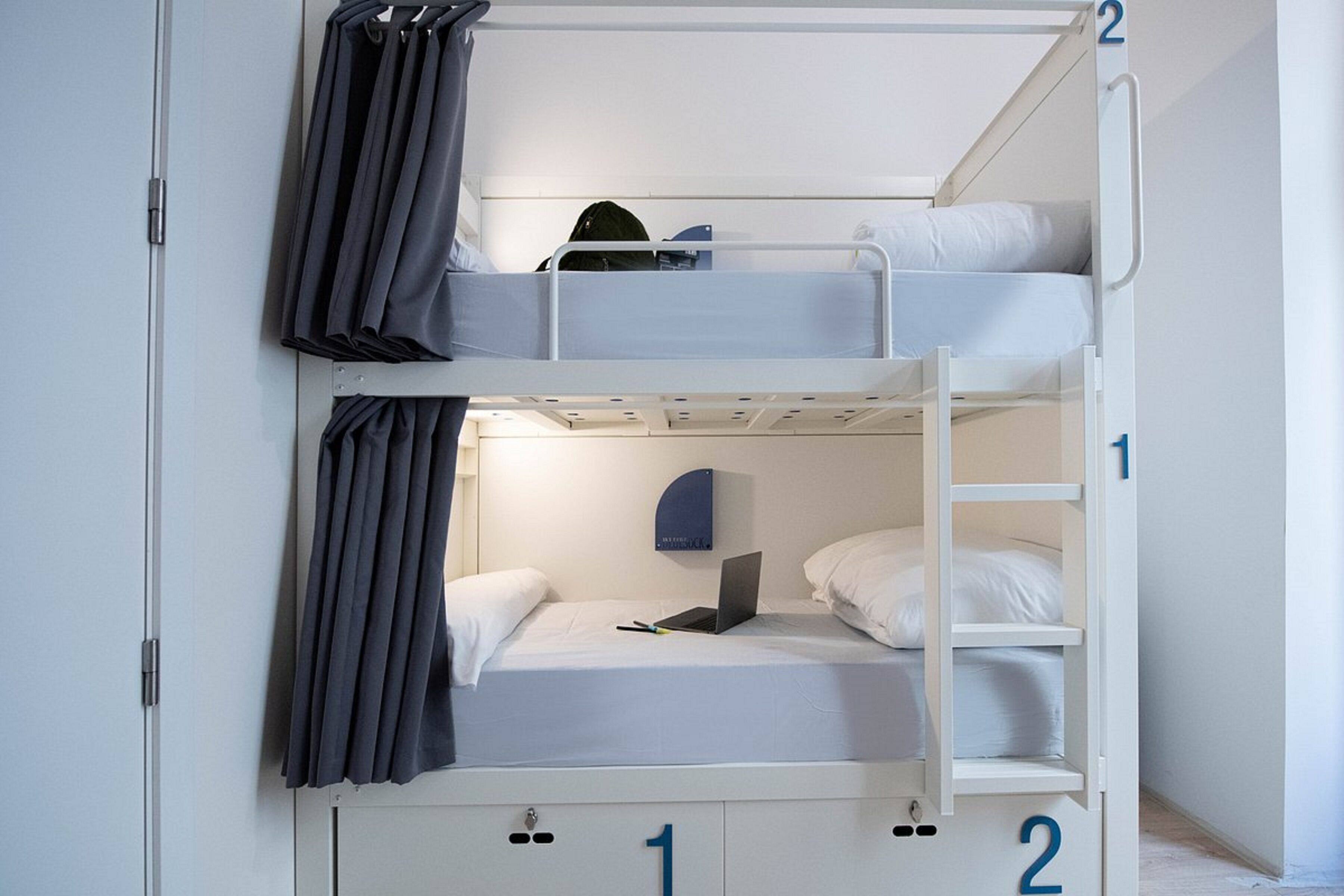 Een slaapkamer met stapelbed, een laptop op het onderste bed, een deur met een nummer en een gordijn aan de linkerkant.