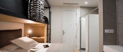 Una habitación de hotel moderna con una cama litera, una mesita de noche con lámpara, un estante pequeño y un área de ducha.