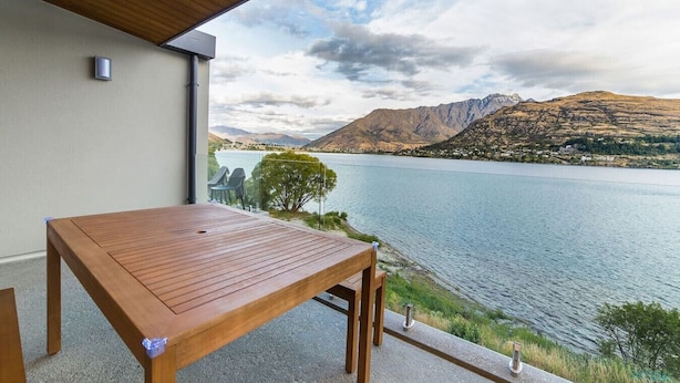 Una mesa y sillas de madera en un balcón con vistas a un lago y montañas al fondo.