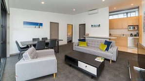 Una sala moderna con un sofá gris, una mesa de centro y un comedor con mesa y sillas. Se observa una zona de cocina con armarios y electrodomésticos.