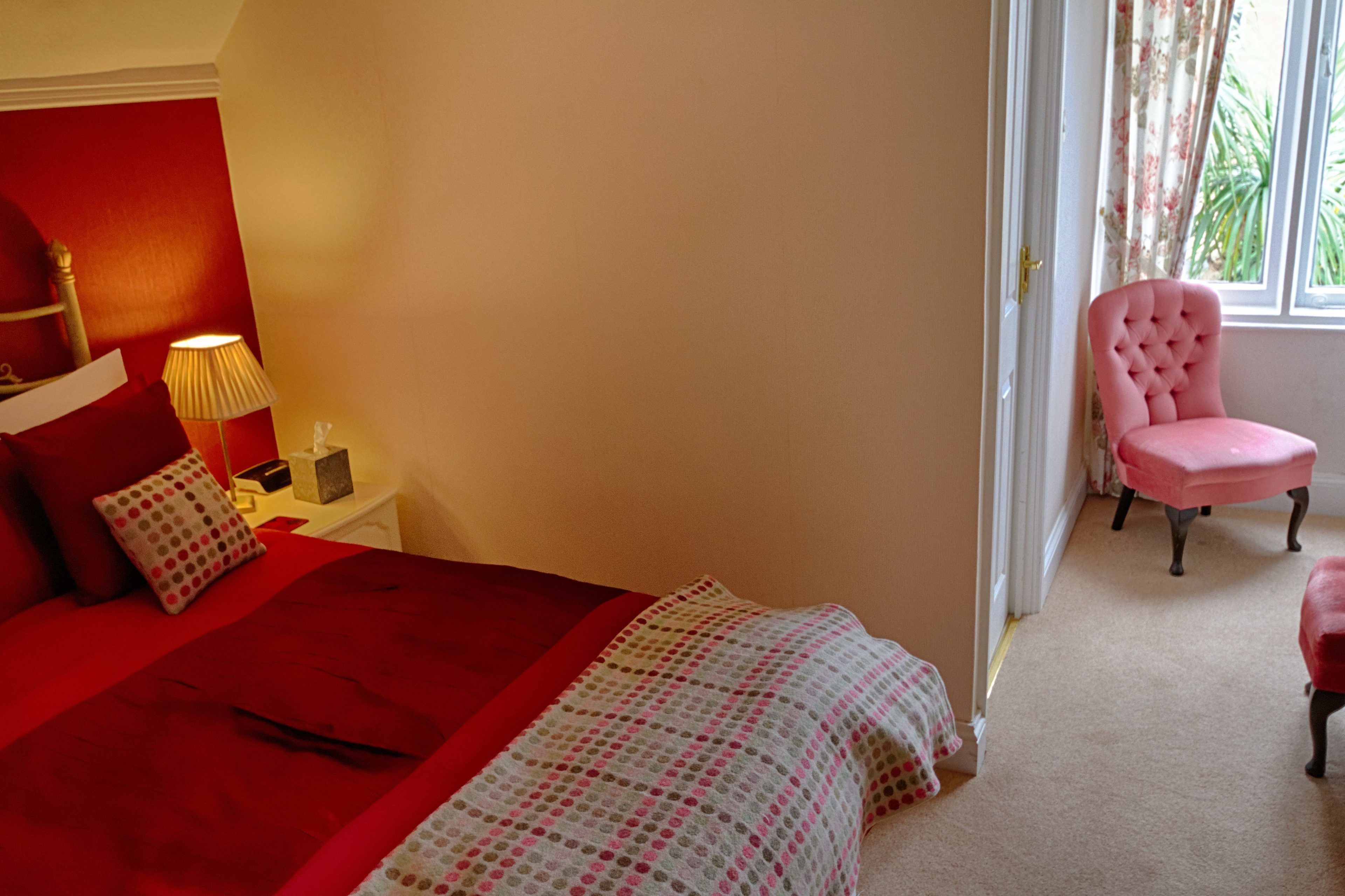 A bedroom with a red bedspread, a pink chair, and a nightstand with a lamp.